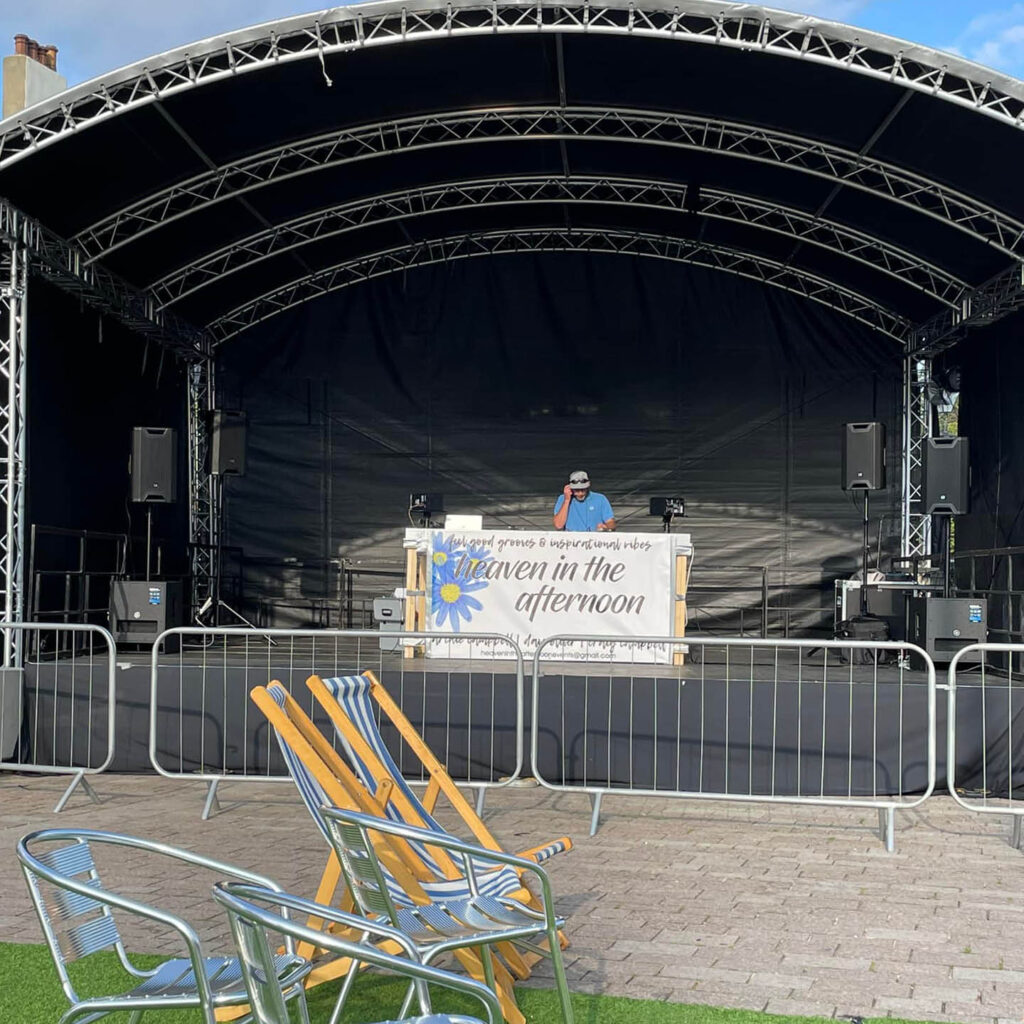 A DJ stands behind his deck on a large stage. A white sign with a blue flower is attached to the front of the deck. It says Heaven in the Afternoon.