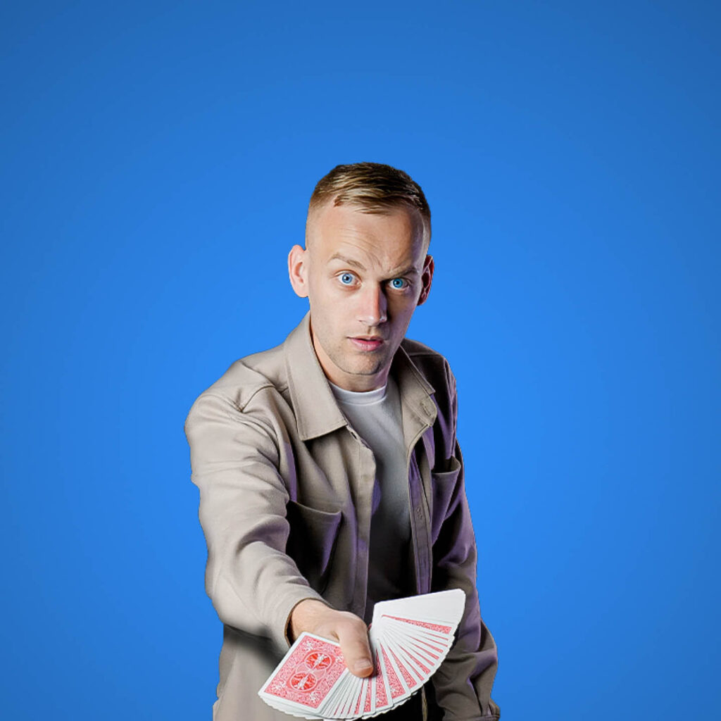 Jack Rhodes is a white man with short blonde hair. He wears a beige jacket over a white t-shirt and stands in front of an entirely blue background. He raises his eyebrows daringly as he holds out a fanned set of cards.