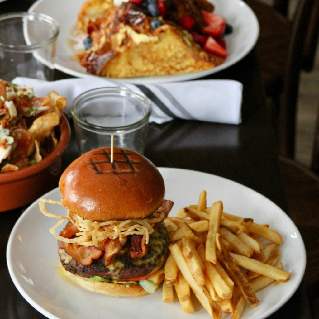 A stacked burger sits on a white plate. It is filled with what looks like a burger patty, bacon and crispy onions. Golden fries sit on the plate next to it. In the background, a plate of french toast and berries is visible.