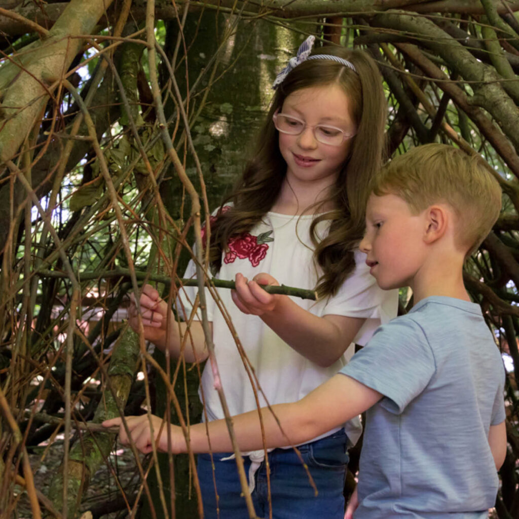 A young boy and girl work together to weave sticks together to create their den in the woods.