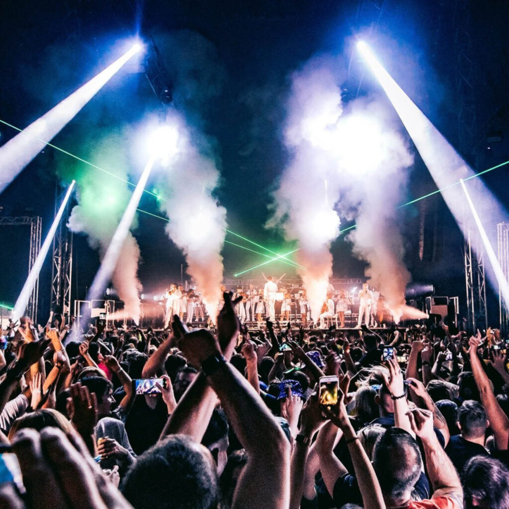A huge crowd of people raise their hands and jump in front of a smoking stage. Strobe lights beam down on the crowd.