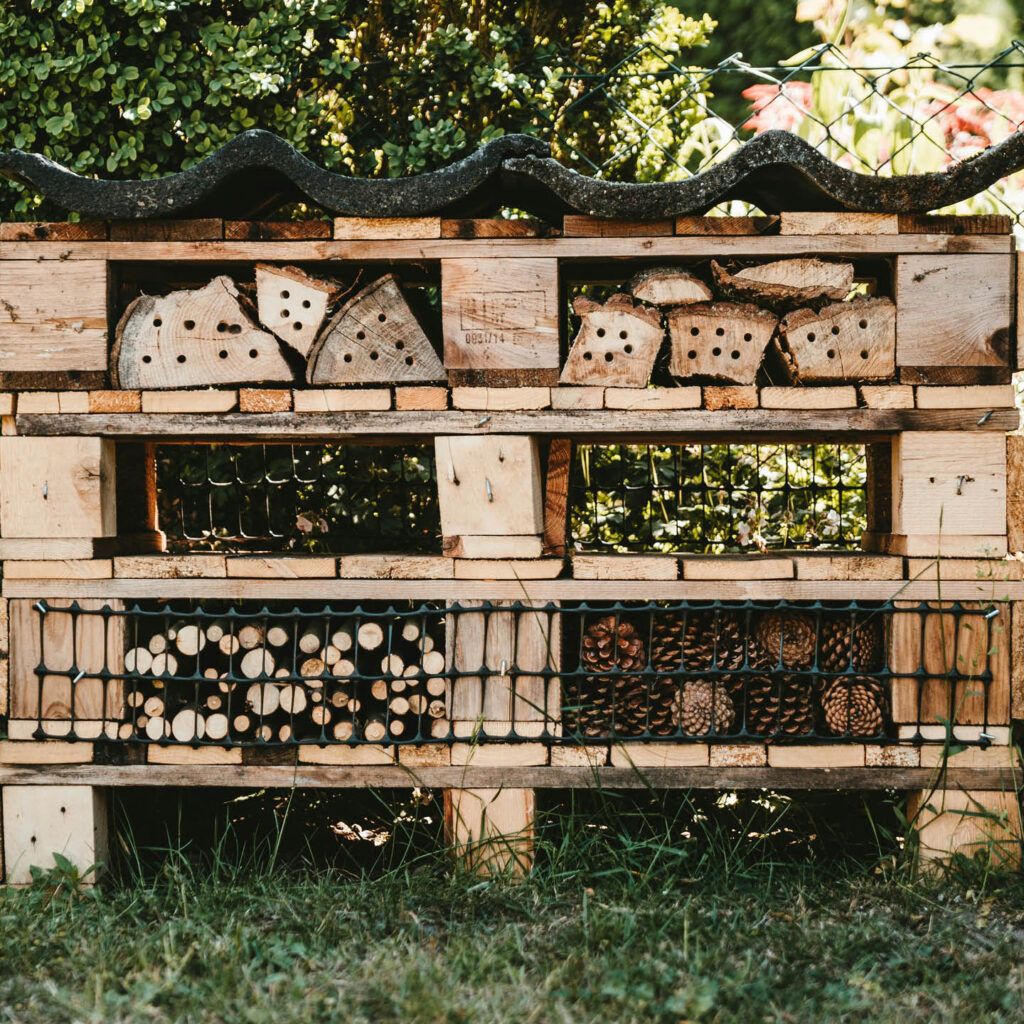Three wooden pallets are stacked on top of each other and filled with wood, sticks, tubes and other materials. It is topped with a corrugated roof. It is a large insect hotel for the garden.