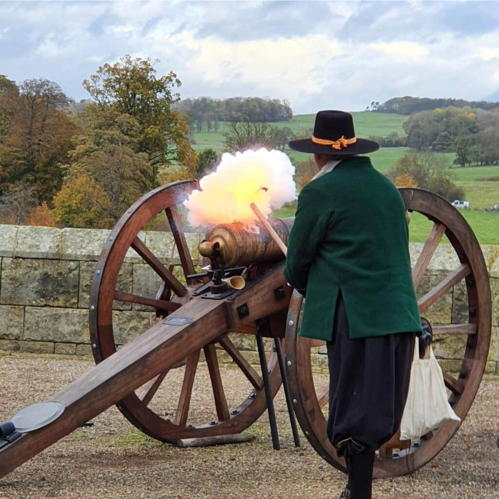 A man dressed in a green coat and black hat leans over a wooden cannon in the grounds of Alnwick Castle. Smoke and fire plume as he lights it.