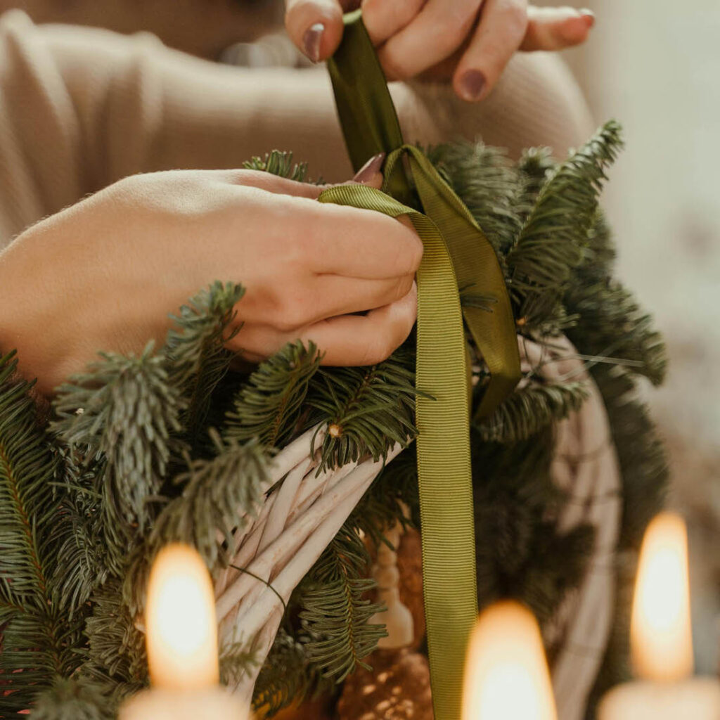 A person ties a green ribbon to a Christmas wreath. Candle flames glow in the foreground.