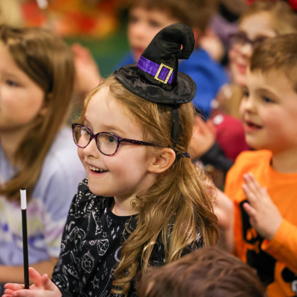 A little girl dressed as a witch sits in a crowd of other children. She smiles in awe at something she sees, holding her magic wand between her hands.