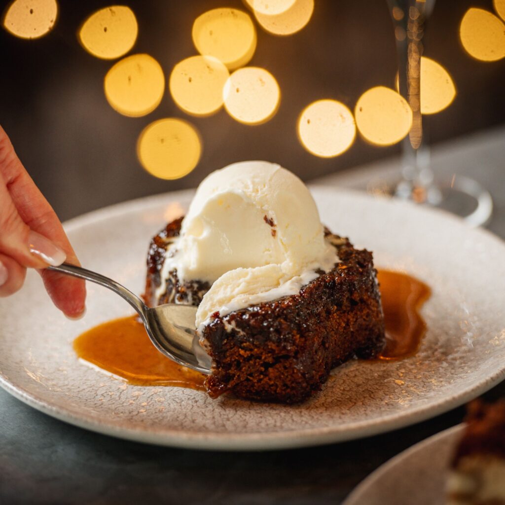 A woman's hand uses a spoon to tuck into a delicious dessert of sticky toffee pudding