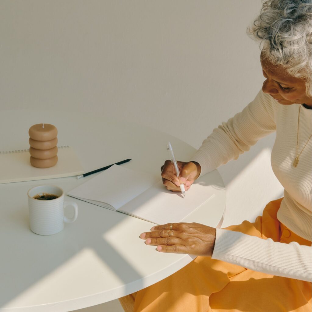 A woman is seated at a talle with a cup of coffee, journalling in her notebook