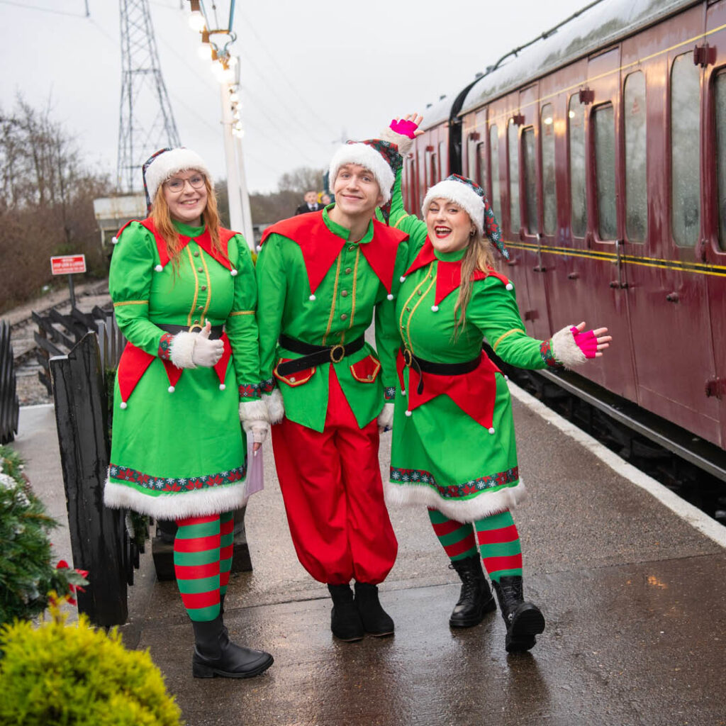Three people dressed as elves smile at the camera on the platform of a train station. Next to them is a old-fashioned maroon train.