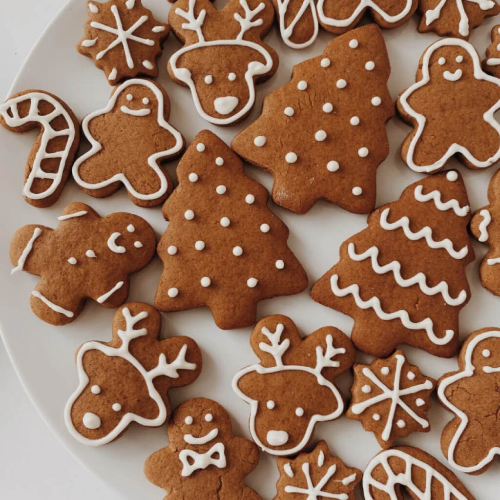 A white plate covered in different gingerbread shapes, including gingerbread people, reindeer faces and snowflakes. They are iced with neat white lines and dots.