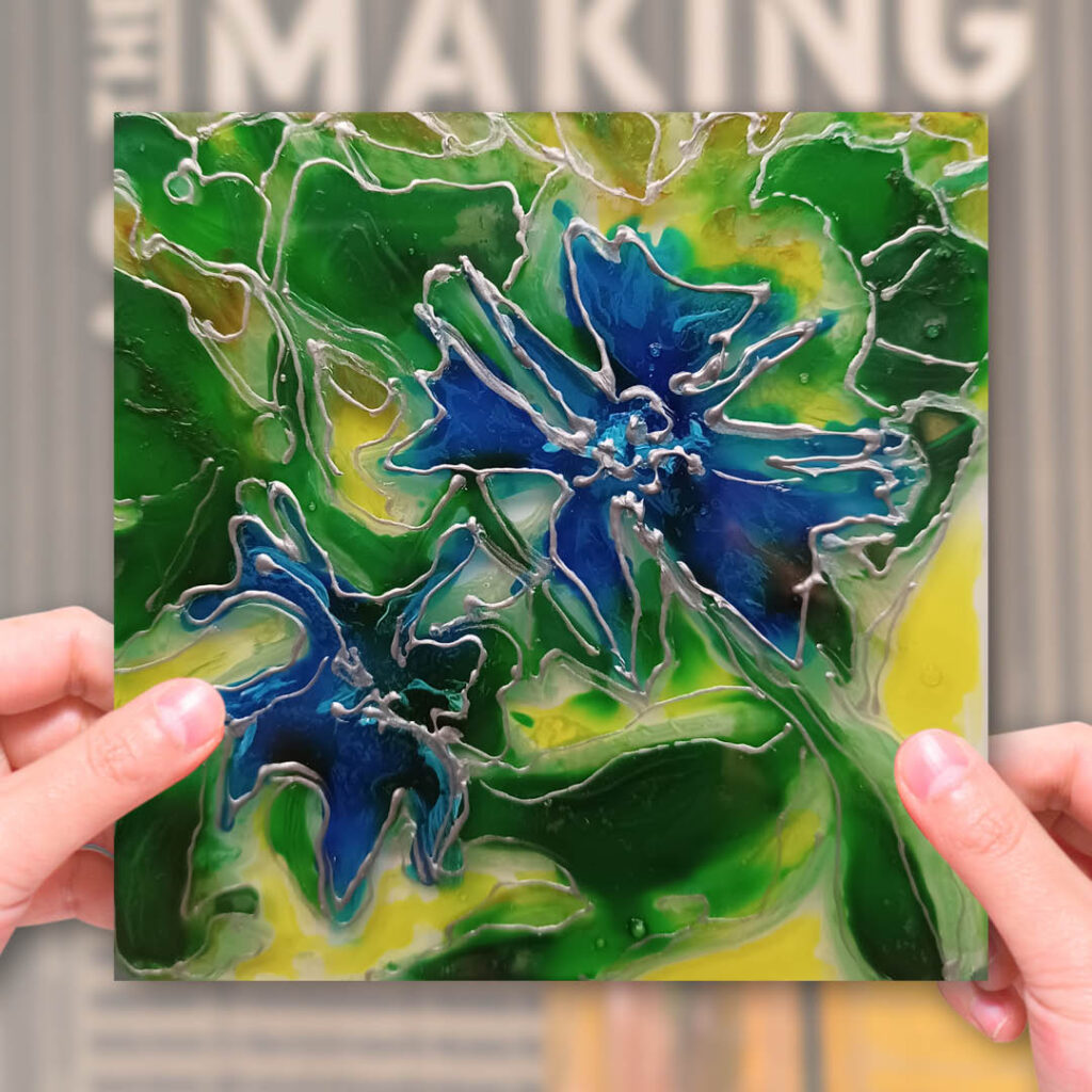 A person holds up a glass tile featuring a green and blue flower and leaf design.