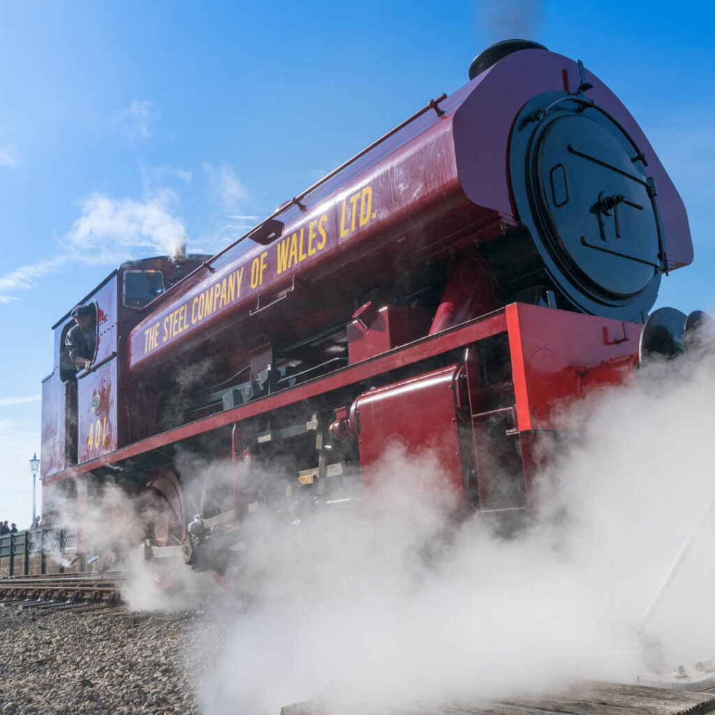 A large red steam train pumps out steam against a bright blue sky.