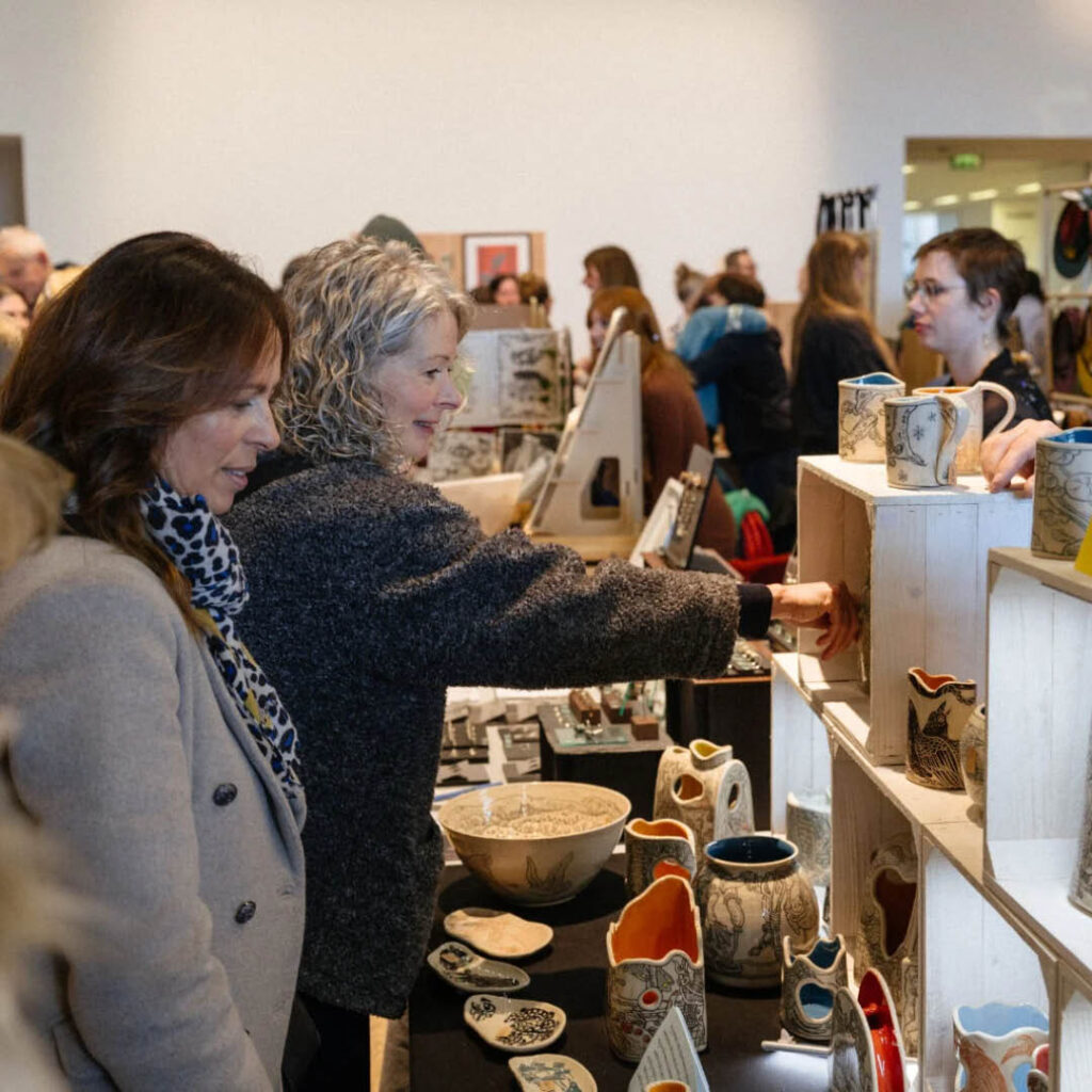 Two women browse a stall selling handmade ceramics. In the background, more people walk around the other stalls at the Winter Makers Market.