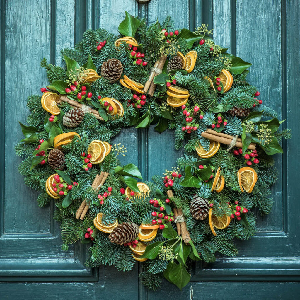 A Christmas wreath hangs on a blue door. It is made from pine leaves, dried orange, cinnamon sticks and berries.