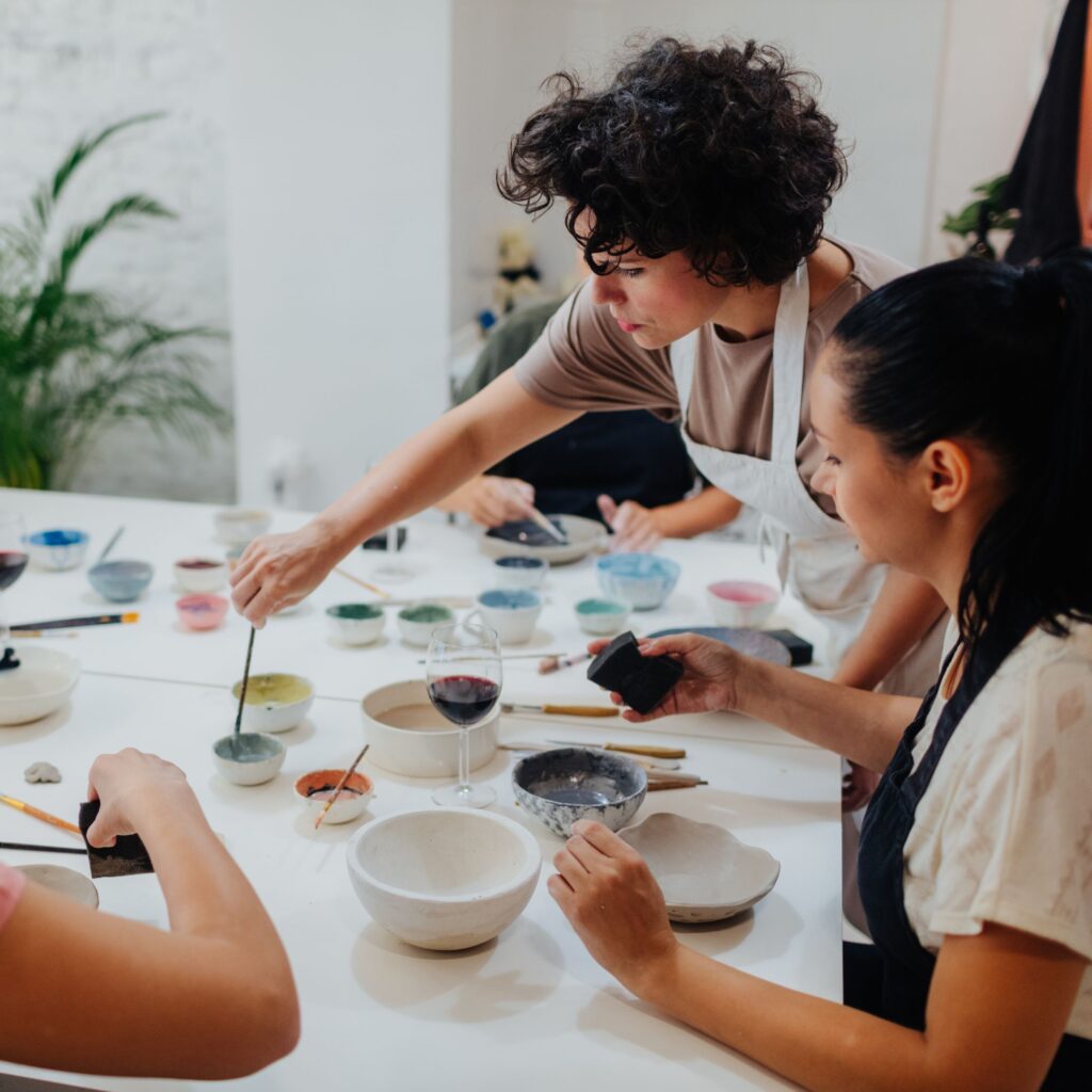 A painting class is taking place around a big square table. Two woman sit next to each other one holds a glass of wine and the other reaches to dip her paintbrush into paint.