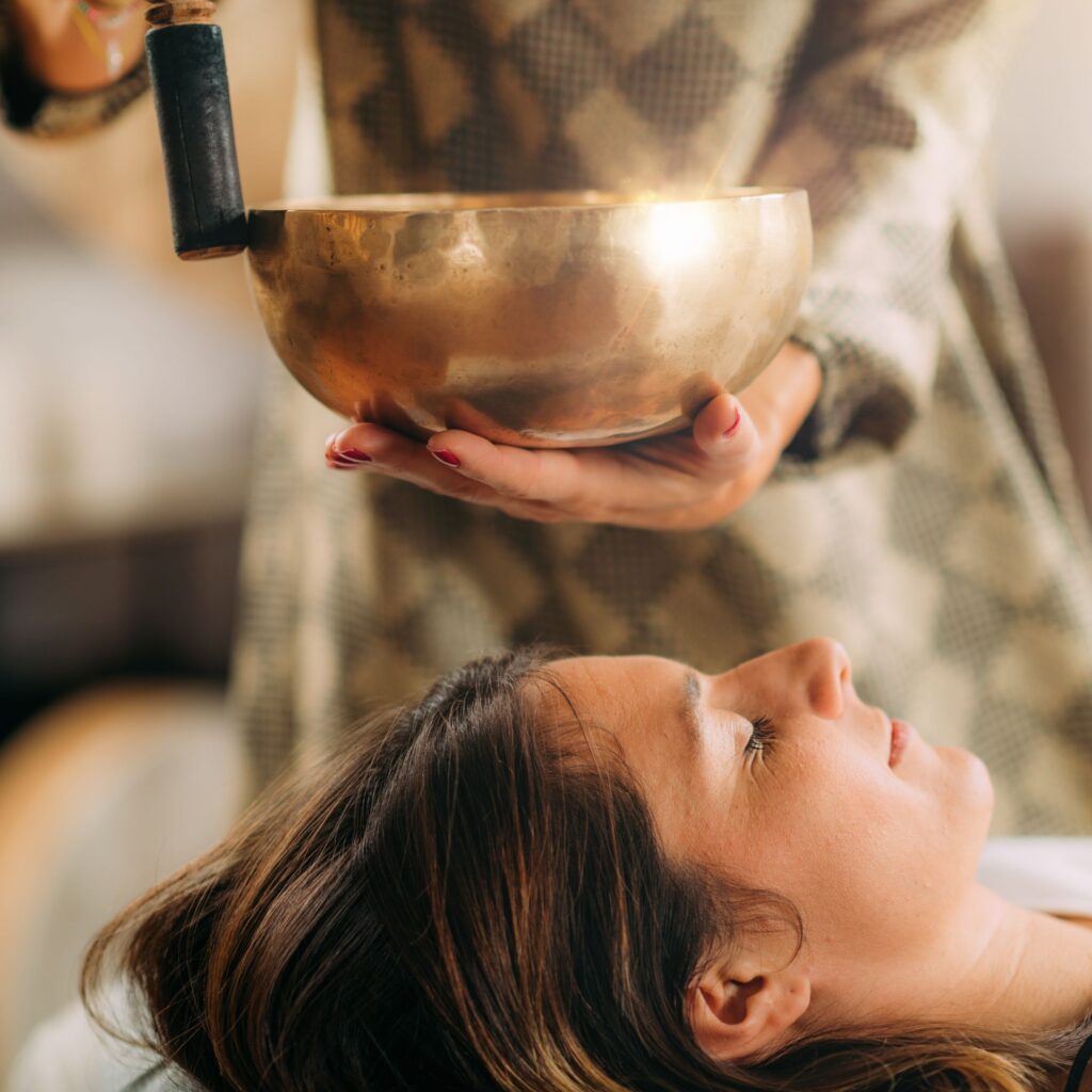 A woman lays on her back with her eyes closed as someone plays a sound bowl near her head to calm the mind.