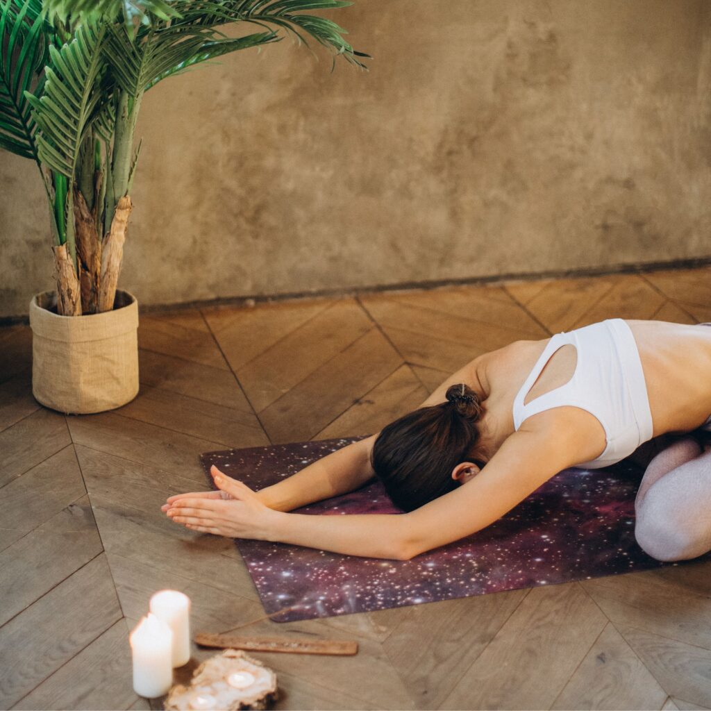 In a yoga studio a woman is lying in childs pose on a purple yoga mat next to a plant and some candles. She has her hair in a low bun and wears a white sports bra.