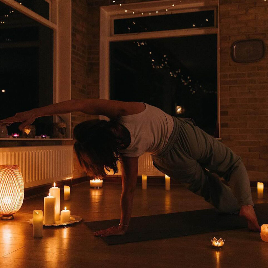A woman with her back to the camera does a side plank on a yoga mat. She stretches her arm above her head. The studio is dimly lit with candles scattered around the floor.