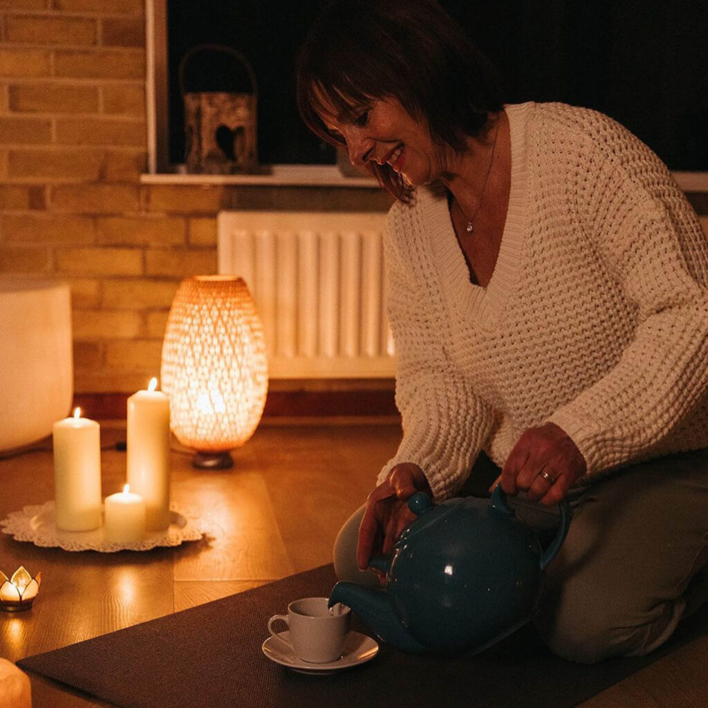 A woman smiles as she kneels on the floor pouring tea into a teacup. The room around her is dimly lit with a warm lamp and candles.