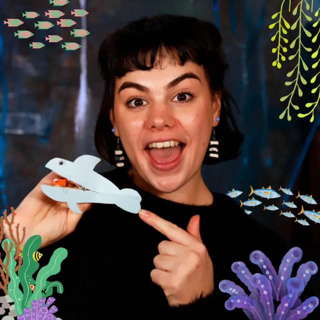 A woman opens her mouth in amazement as she holds up a small blue paper fish with an open mouth.