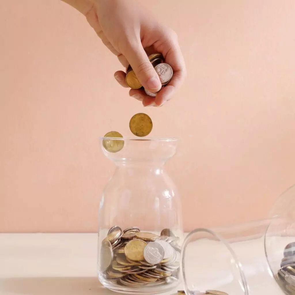 Piggy bank, a hand drops loose change and coins into a clear jar to save money