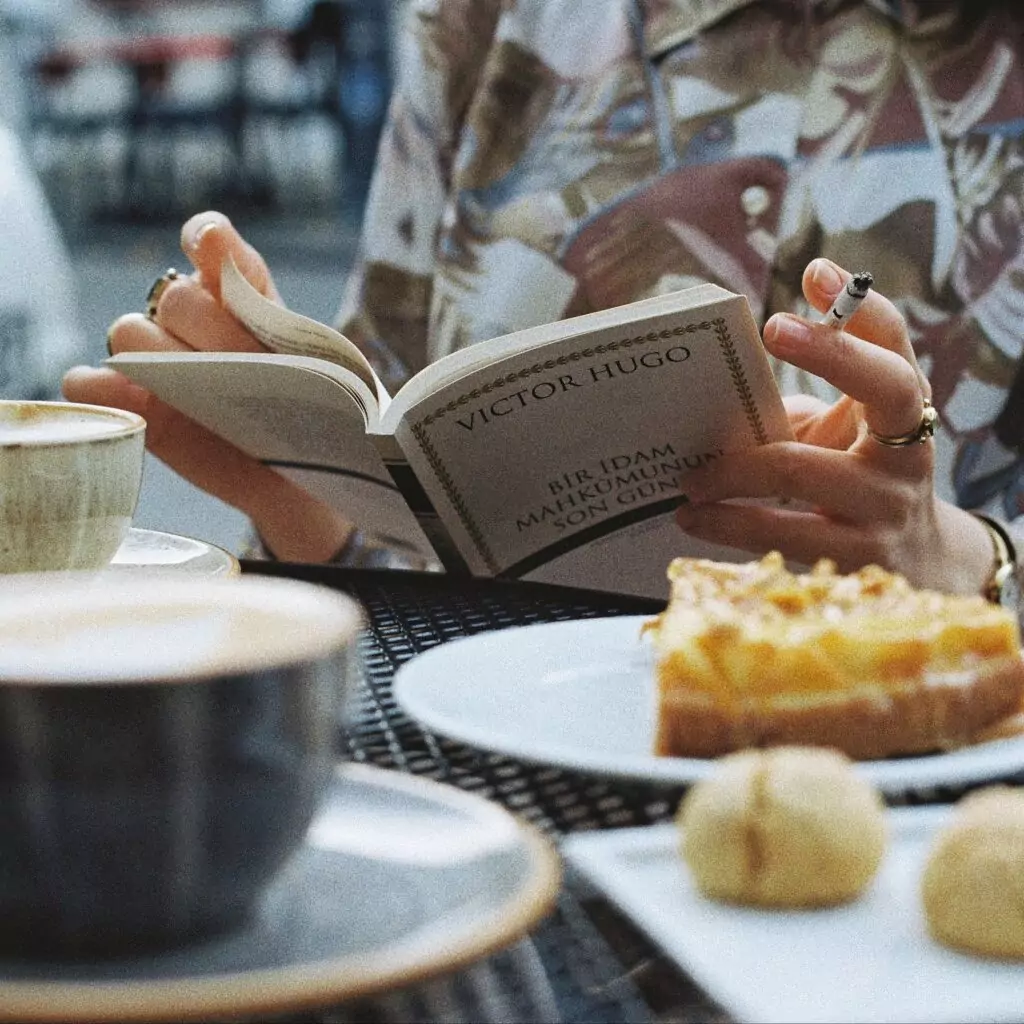 At breakfast a women has a cigarette while reading her book and drinking coffee