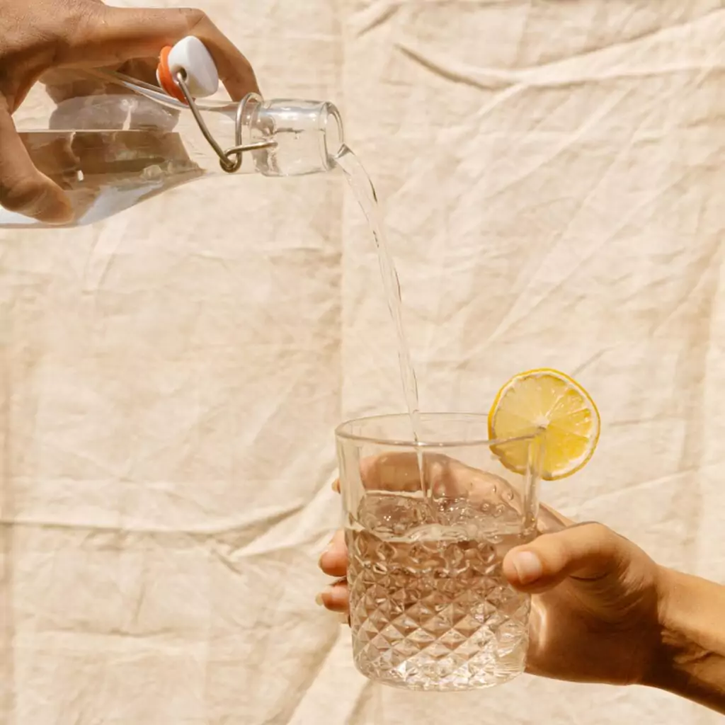 One hand holds a clear glass filled with water and a side of lemon while a second hand pours from a jug of water