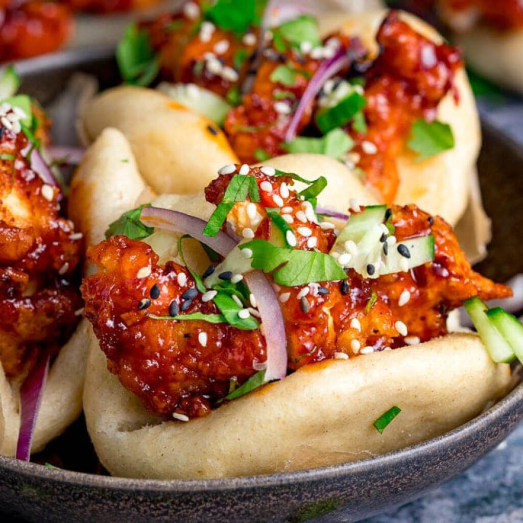 A plate of fresh bao buns filled with crispy marinaded chicken, onions, sesame seeds and coriander garnish.