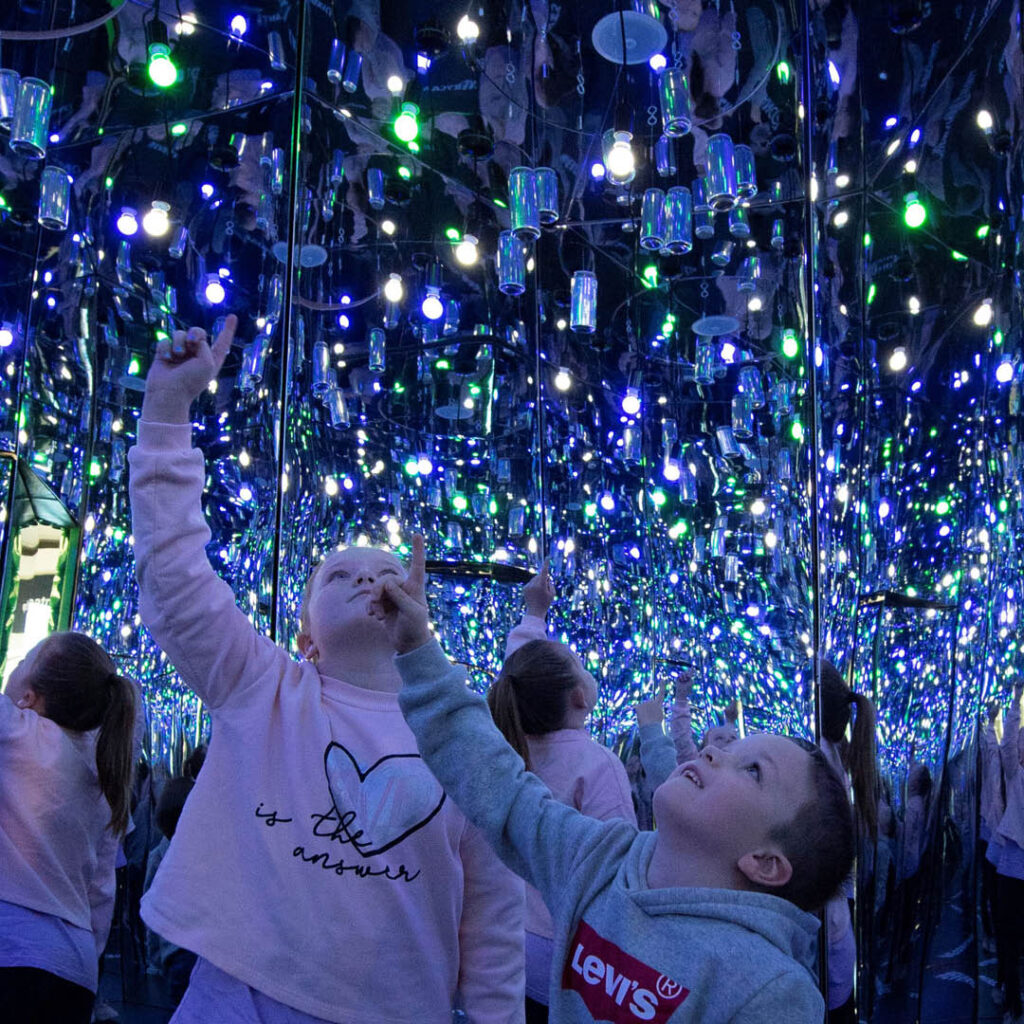 Two young children point and gaze upwards in wonder as they stand inside a mirrored infinity room with hanging aluminium cans. It is an exhibition at the Life Science Centre this February half term.