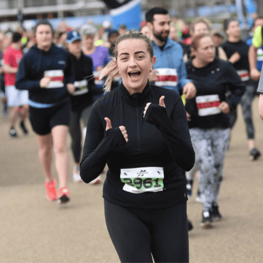 A young woman grins and gives a thumbs up to the camera as she runs the Newcastle 10k race. Other runners can be seen behind her.