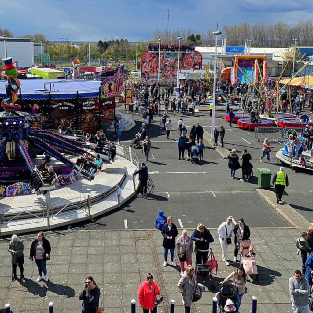 Different funfair rides and stalls are scattered across a large car park at Newcastle Quays. People mill about between them.
