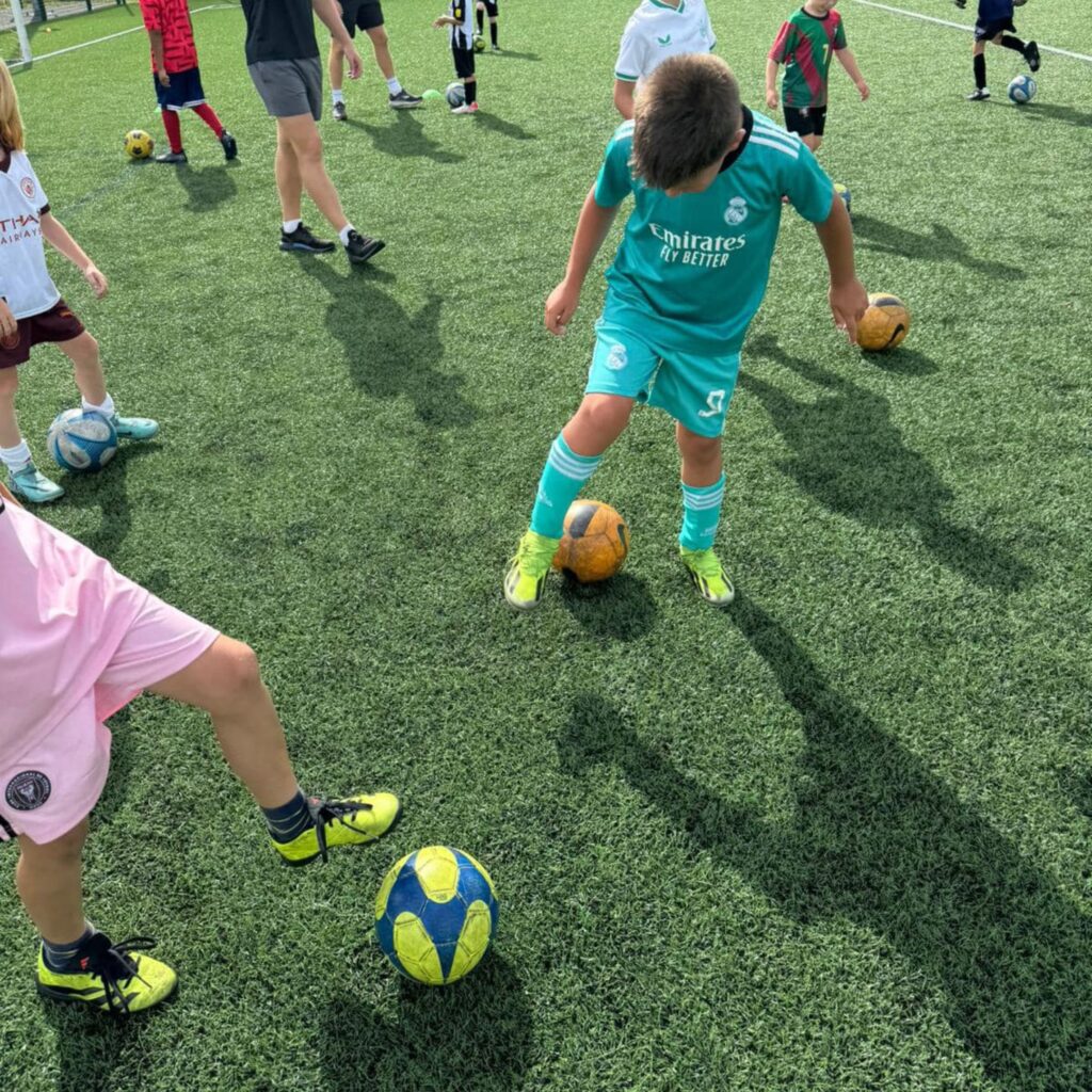 Young boys kick around footballs on a green field.