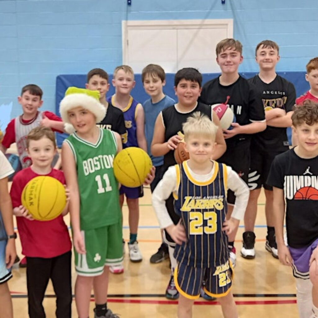 Young children in basketball bibs stand in a gym. Some of them hold basket balls. They grin at the camera.