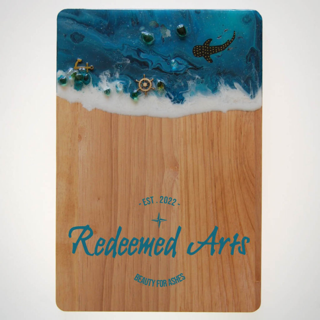 A wooden chopping board features a deep blue acrylic design on one end, which looks like ocean waves. Small sea creature embellishments have been added to the paint.