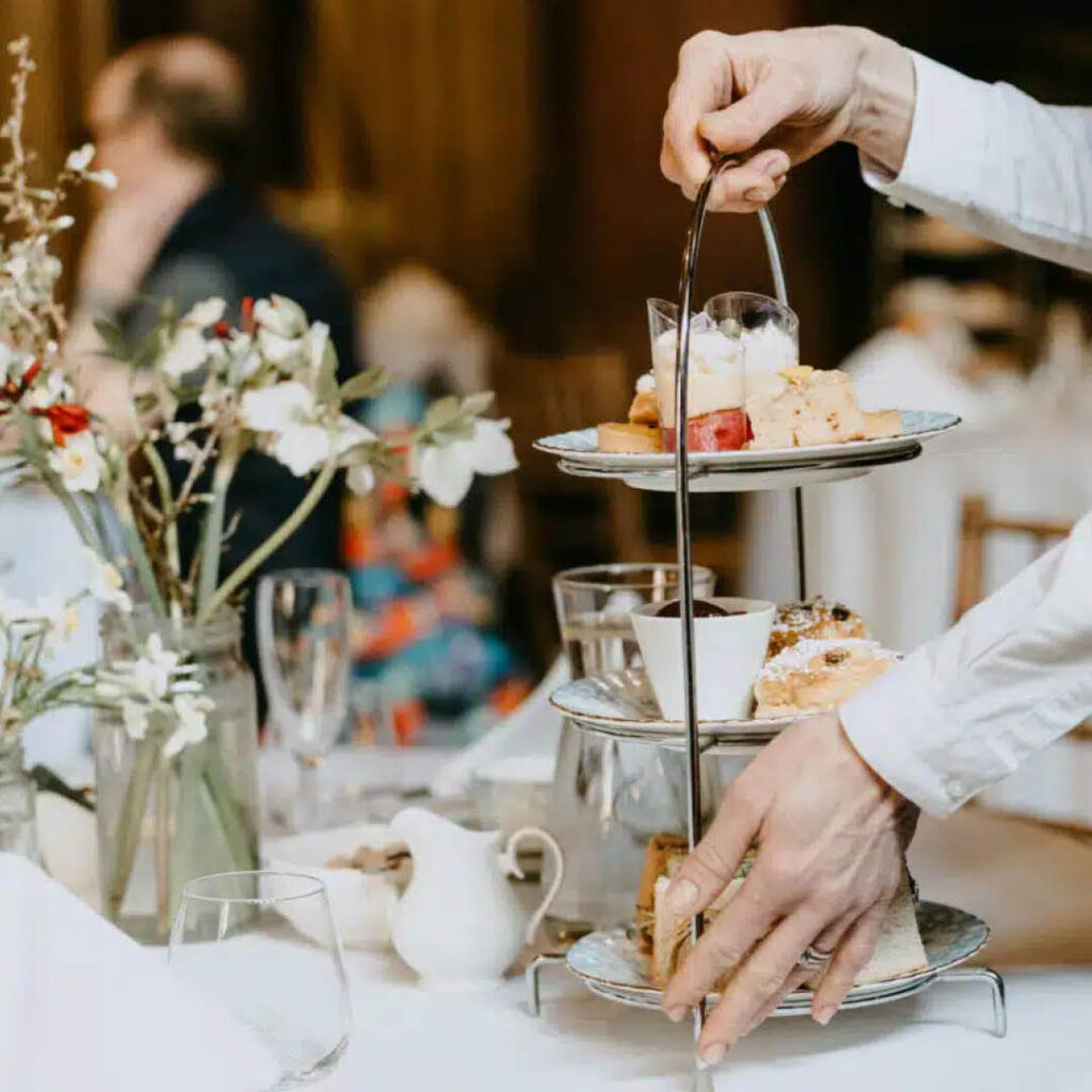 An unseen waiter places a three-tier afternoon tea platter onto a white tablecloth. Delicate flowers in a clear vase stand nearby.