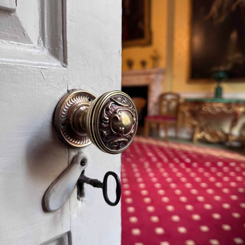 A white wooden door with an intricate brass door knob and key opens onto a room with red carpet and ornate furniture. Guests can visit rooms like this on a Behind The Scenes Tour of Raby Castle.