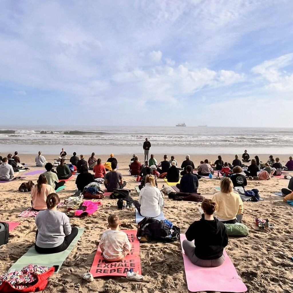 Lots of people sit on yoga mats on a sandy beach. They face the sea, where an instructor stands ready to lead them through a yoga flow.