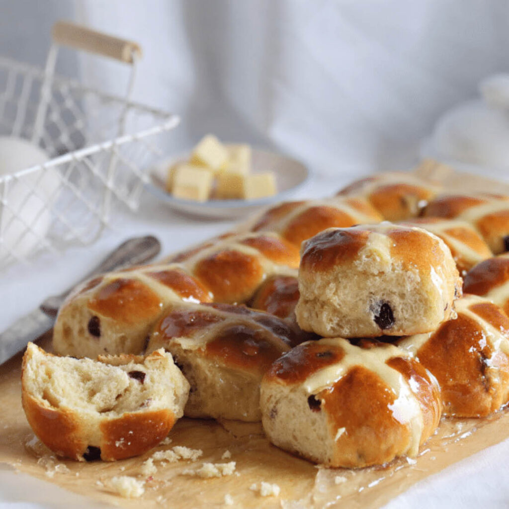 A freshly baked batch of glazed hot cross buns sit on a wooden board. One of them has been torn open, ready to be buttered.