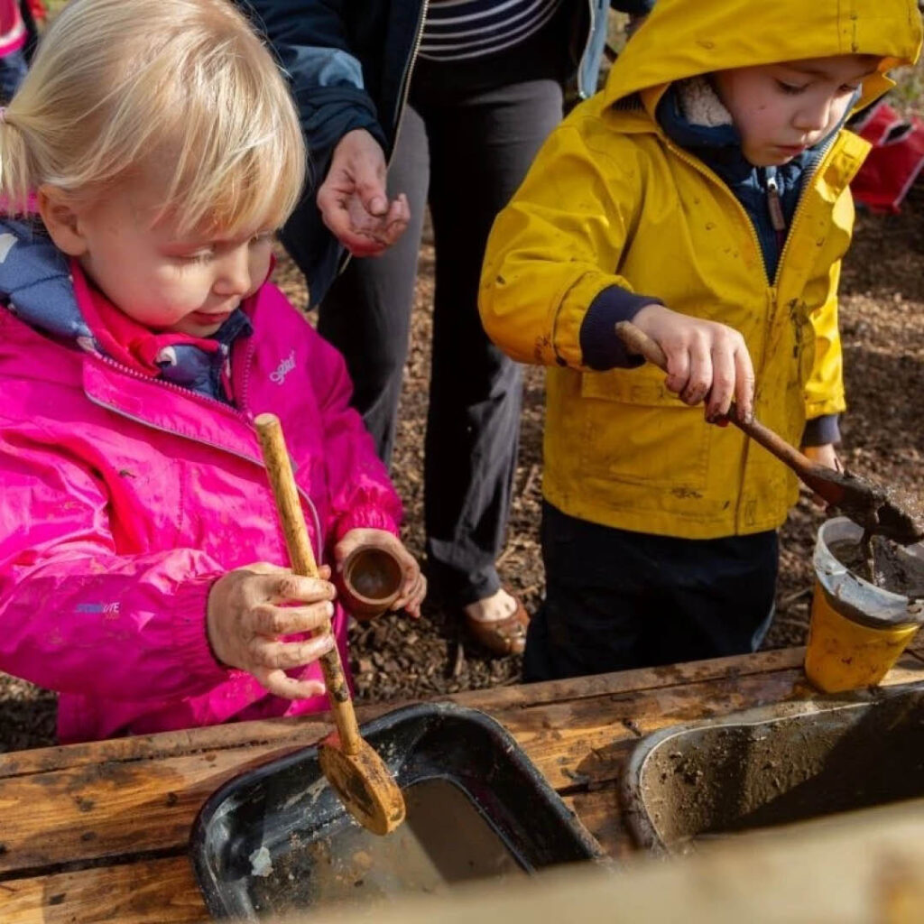 Two young children in yellow and pink raincoats use small shovels to scoop mud out of a trough. They are enjoying Mudfest at Washington Wetland Centre.