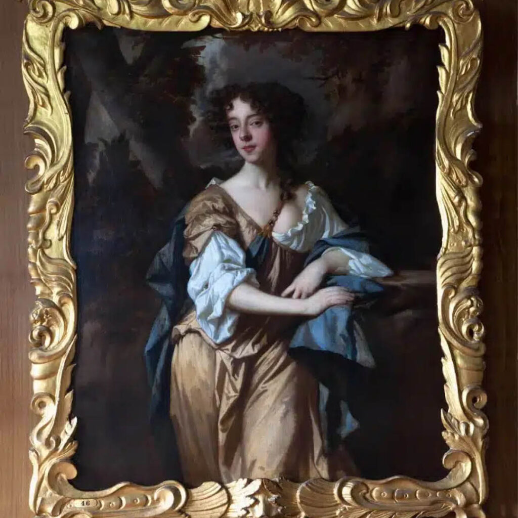 A gold framed portrait of a young woman with brown curly hair. She wears a long brown dress with puffy blue sleeves and tilts her head the the side as she looks towards her painter. This is one of the portraits of women on display at Raby Castle.