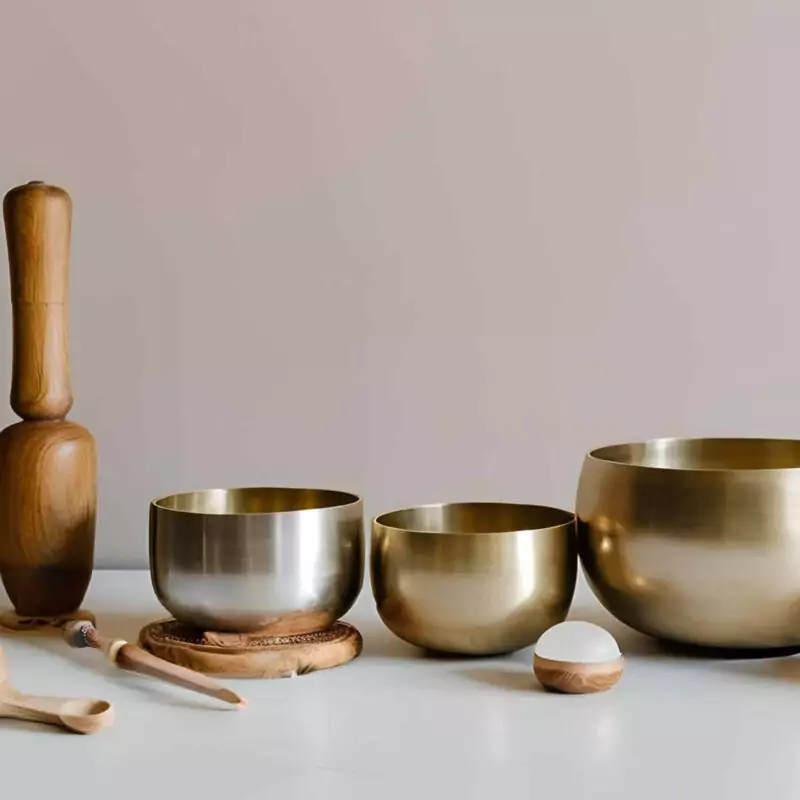 Three gold metal singing bowls of different sizes are lined up on a white surface. There are also wooden batons and sticks for creating sounds on the bowls.