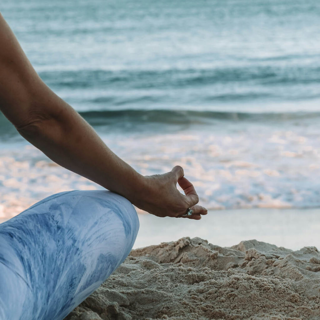 A person sits cross legged on the sandy shoreline, facing the sea. They press their thumb and index finger together in a meditative position.