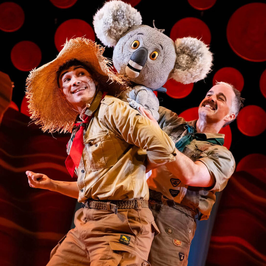 A person dressed in a straw hat and brown explorers outfit grins as they look over their shoulder at the koala puppet on their back. Another man smiles as he holds the koala in place. They are performers in the stage show: The Koala Who Could.