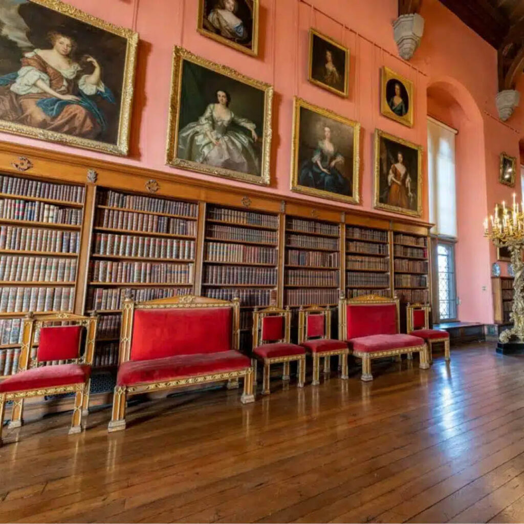 Huge portraits of women in large dresses line the pink wall above a wall-length bookshelf in Raby Castle.