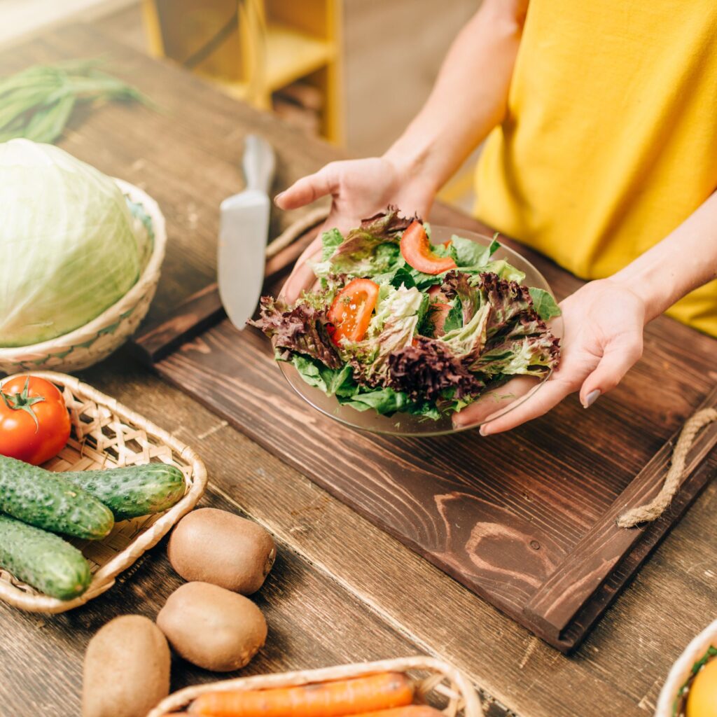 A woman out of frame holds a bowl of salad above a cutting board and fresh vegetables