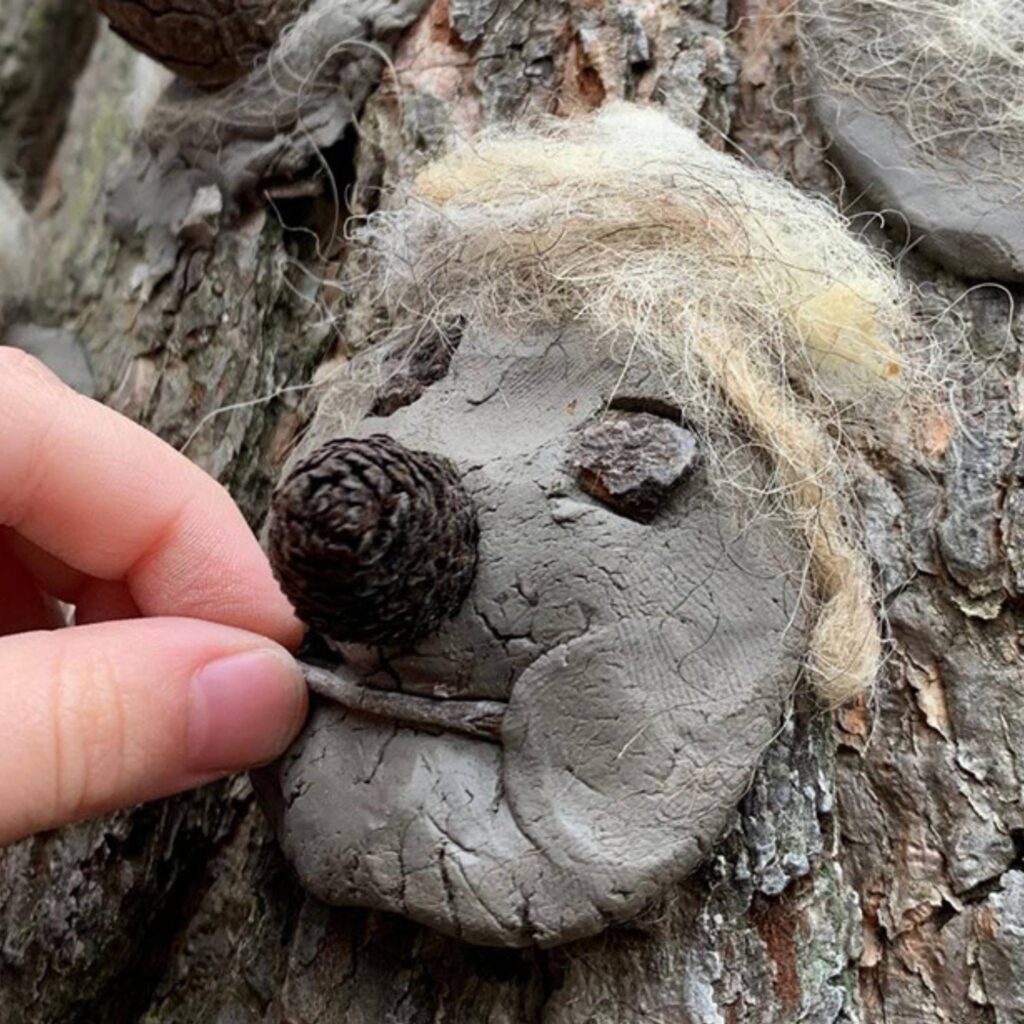 An unseen person places a stick onto a flattened piece of clay on a tree trunk. It is a clay face with a pinecone for a nose, stones for eyes and wool for hair.