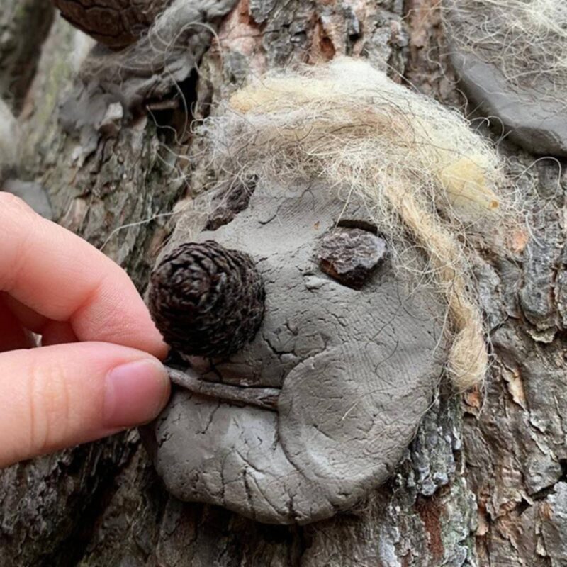 An unseen person places a stick onto a flattened piece of clay on a tree trunk. It is a clay face with a pinecone for a nose, stones for eyes and wool for hair.