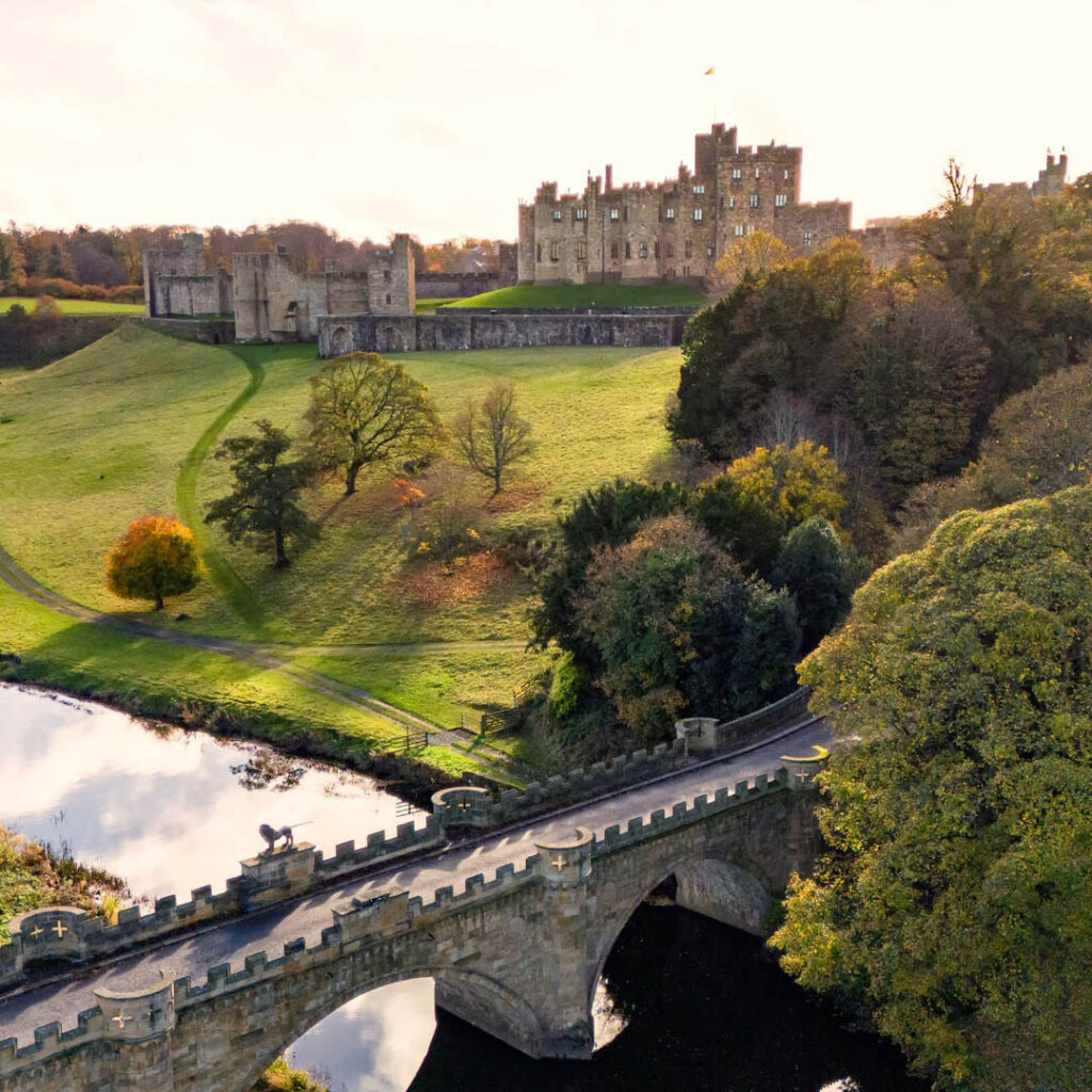 Alnwick Castle perches on a green hill, which slopes down to meet a river and stone bridge with a cluster of trees. The afternoon sun shines down.