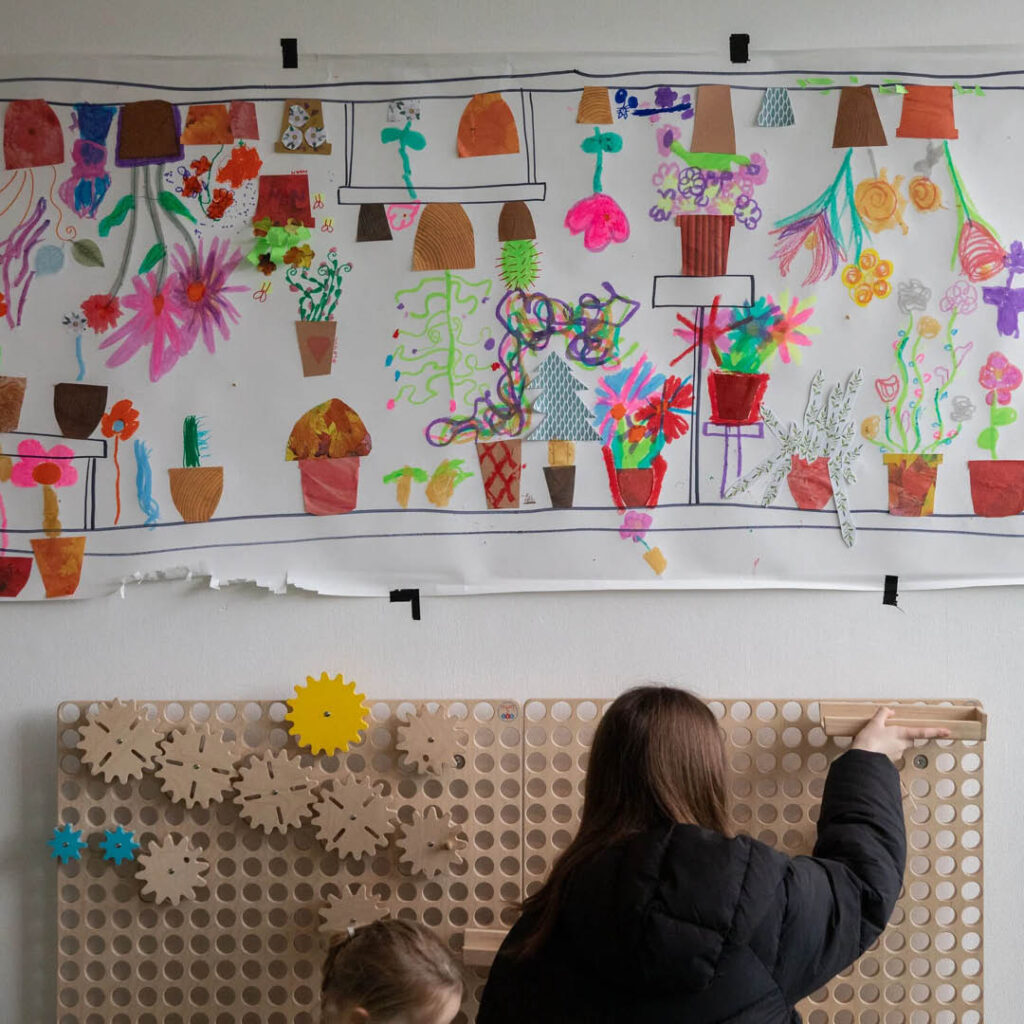 Two girls fix wooden parts to a wooden board with holes in it. Above them, there is a wall of artwork featuring flowers and plant pots created by kids.