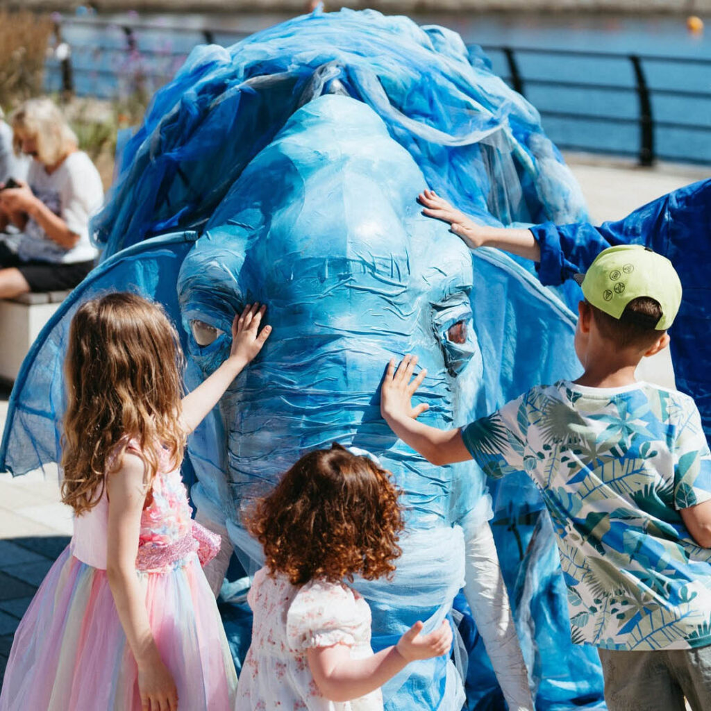 Three young children stroke the trunk of a huge blue elephant puppet in the sunshine.