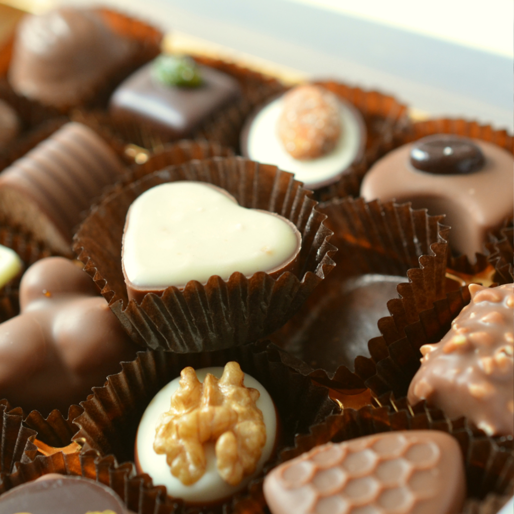 A box of variously coloured Belgian chocolates. Some are topped with nuts and honeycomb shapes. A white heart shaped one sits on top of them all at the centre.